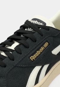 Gros plan d'une basket Reebok noire et blanche montrant des lacets noirs, une semelle beige et un logo Reebok doré sur le côté.