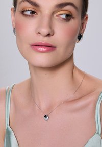 Srebrny naszyjnik z okrągłym zawieszeniem z czarno-białymi akcentami. Modelka ma na sobie dopasowane okrągłe kolczyki wkręty oraz miętowozieloną bluzkę.