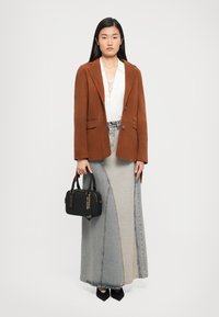 Woman wearing a brown blazer, white lace-up top, long patchwork denim skirt, black pointed shoes, holding a black handbag with gold accents.