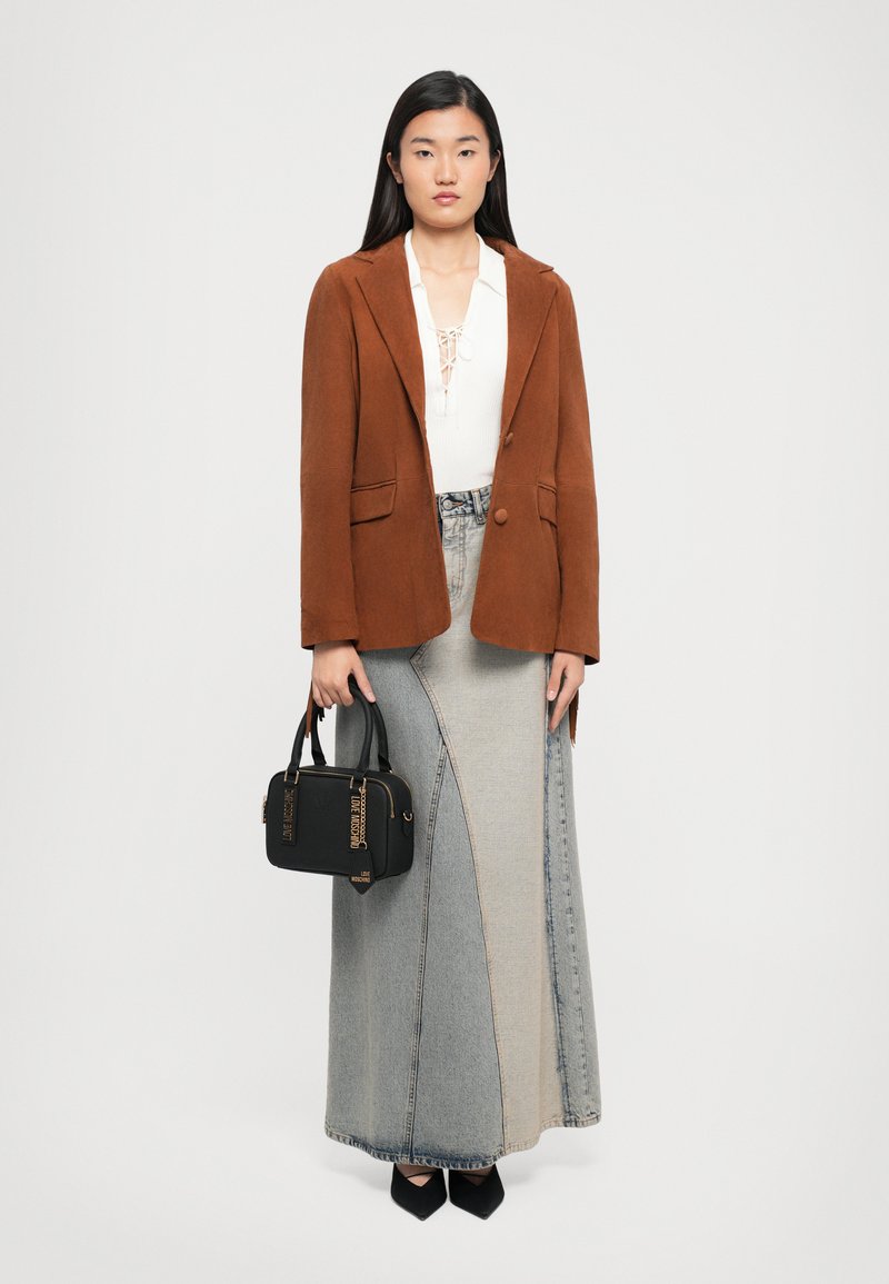 Woman wearing a brown blazer, white lace-up top, long patchwork denim skirt, black pointed shoes, holding a black handbag with gold accents.