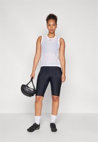 White mesh athletic tank top with black cycling shorts; person holds a black bicycle helmet and wears black cycling shoes and white socks.