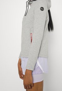 Veste en polaire grise avec capuche, poches zippées, accents de fermeture éclair rose et cordons de serrage noirs. Présente un logo rond sur la manche.