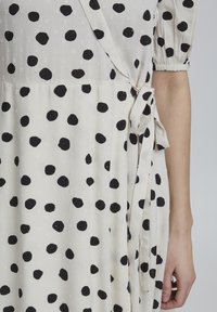 Vestido blanco con lunares negros, que presenta un diseño cruzado, mangas cortas abullonadas y un detalle de cinturón atado en la cintura. Textura de tela suave.