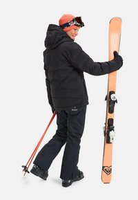 Zwarte pufferjack, zwarte skibroek en zwarte handschoenen. Houdt een oranje ski vast met zwarte bindingen en rode skistokken, met een roze beanie en goggles.