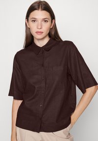Camisa de botão de manga curta castanha, feita em linho, com colarinho, um bolso no peito e um corte descontraído. Superfície texturada.