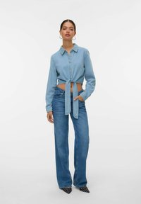 Camicetta in denim blu chiaro, corta, con bottoni e dettaglio a nodo, abbinata a jeans blue a gamba larga e tacchi a punta neri a pois.