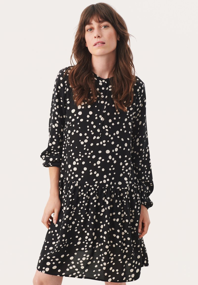 Vestido negro con lunares blancos, mangas largas, escote abotonado y un dobladillo acampanado con un volante fruncido en la parte inferior. Textura de tela suave.