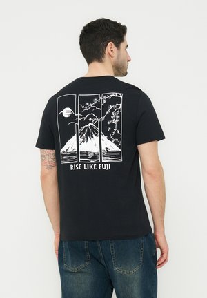 Man wearing black T-shirt with white graphic of Mount Fuji, cherry blossoms, moon, and text "RISE LIKE FUJI" on back, paired with blue jeans.