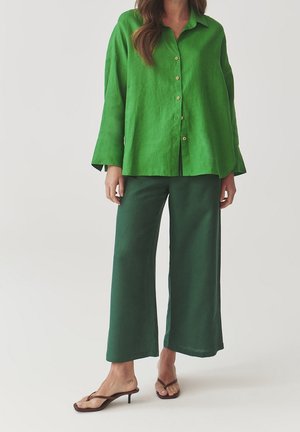 Groen linnen overhemd met lange mouwen, gecombineerd met bijpassende groene brede broek en bruine sandalen met bandjes.