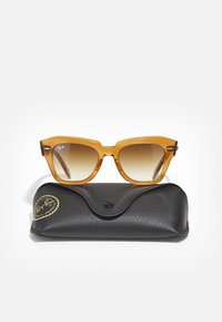 Ochelari de soare din plastic de culoare chihlimbar, cu lentile graduat maro, având o formă clasică de tip cat-eye. Însoțiți de un toc texturat de culoare neagră cu logo.
