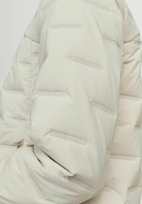 Doudoune beige clair avec un design matelassé, dotée de rayures horizontales perforées et d'une texture lisse et douce. Fermeture éclair sur le côté.