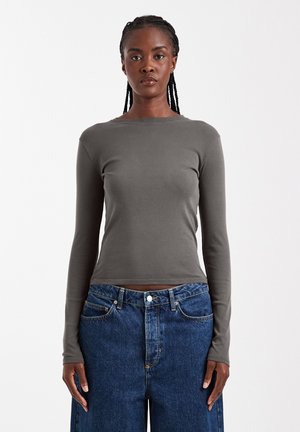 Femme aux cheveux tressés portant un haut gris ajusté à manches longues et un jean bleu ample, debout devant un fond blanc uni.