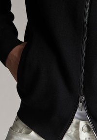 Pull noir à fermeture éclair avec poignets côtelés, doté de poches latérales et d'une texture lisse. La fermeture éclair est visible sur le côté droit.