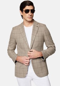 Beżowa marynarka w brązową kratę, teksturowany materiał, klapy typu notch, dwie przednie kieszenie i detal z guzikami. Nosi na sobie białą koszulkę.