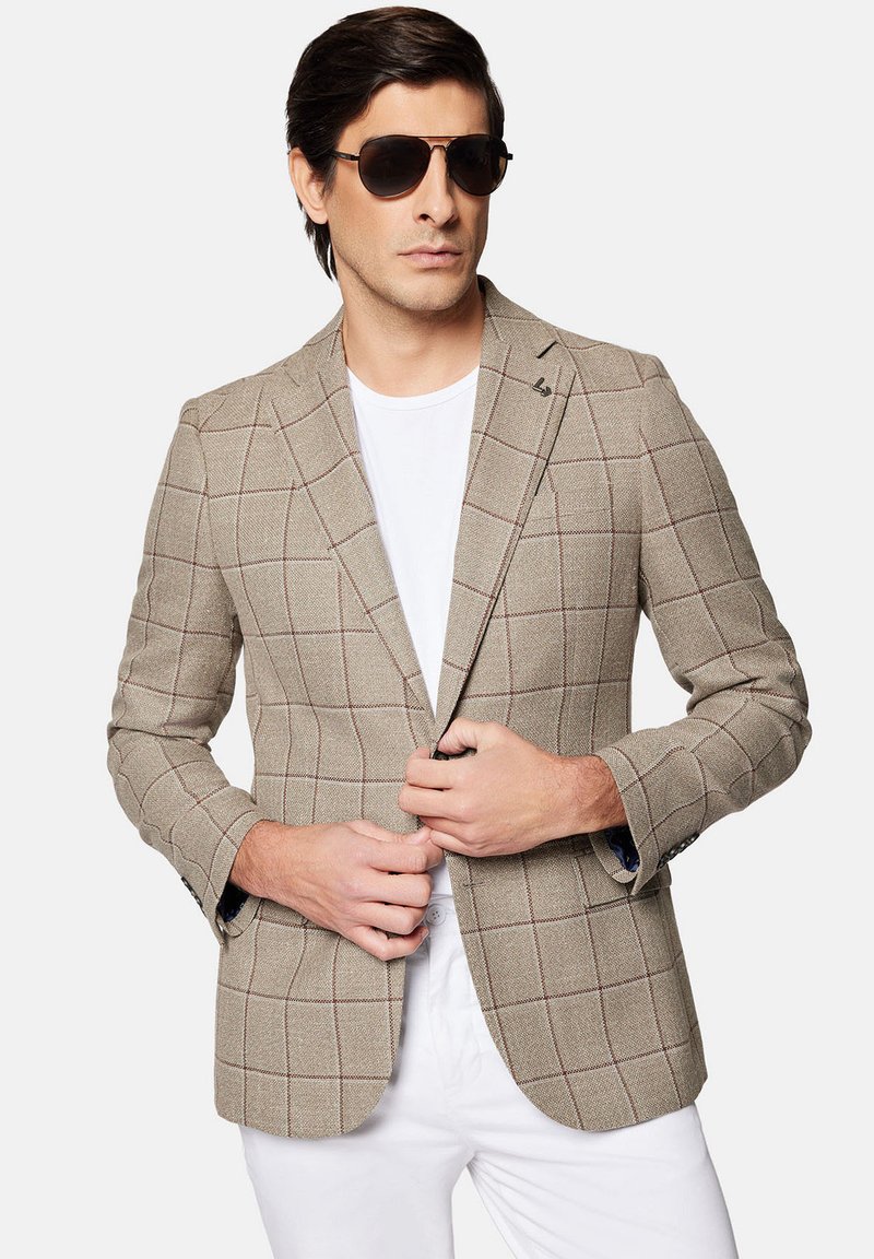 Beżowa marynarka w brązową kratę, teksturowany materiał, klapy typu notch, dwie przednie kieszenie i detal z guzikami. Nosi na sobie białą koszulkę.
