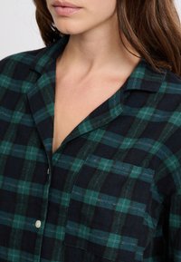 Kobieta nosząca zieloną i granatową koszulę w kratę z guzikami, z kołnierzykiem i kieszonką na piersi, odsłaniająca część szyi i dolnej części twarzy.
