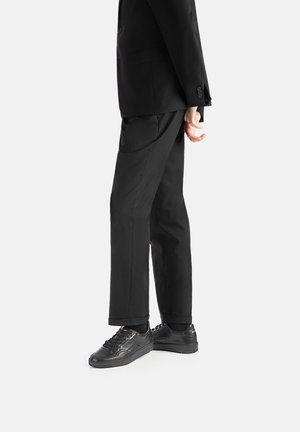 Schwarzer maßgeschneiderter Anzug mit einem Ein-Knopf-Blazer, kombiniert mit schwarzen Sneakern mit strukturierten Mustern und flachen Schnürsenkeln. Glatte Stoffoberfläche.