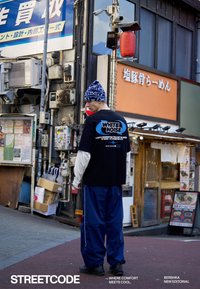 Mladý muž v čiernom tričku s nápisom "White Noise", modrých nohaviciach a vzorovanej čiapke stojí na mestskej ulici pri japonských obchodoch.