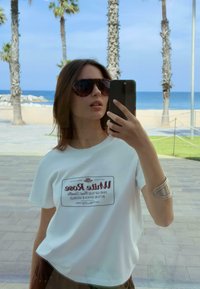 T-shirt blanche avec le texte imprimé "Rose Blanche" et un design graphique. Porté avec des lunettes de soleil surdimensionnées et un bracelet en argent. Cadre réfléchissant.