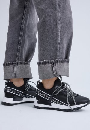 Persona con zapatillas negras y blancas con la marca "exé" y vaqueros grises remangados, de pie sobre un suelo blanco con un fondo gris claro.