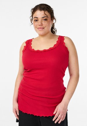 Kiharaliste juuste ja pigmendilaikudega naine, kes kannab punast varrukateta soonikkoes topi pitsiga ääristatud, seisab valge tausta ees.