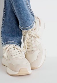 Beige sneakers with white accents, featuring a lace-up design and mixed materials. Paired with blue jeans, showing a casual style.