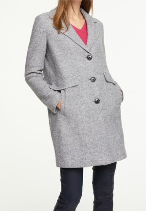 Frau trägt einen grauen Wollmantel mittlerer Länge mit drei schwarzen Knöpfen, seitlichen Taschen und einem roten Oberteil darunter, kombiniert mit schwarzen Hosen.