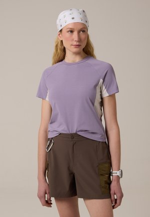 Femme portant un foulard blanc orné de petites abeilles et marguerites, un haut lavande à manches courtes, un short marron avec des poches, une montre et un mousqueton.