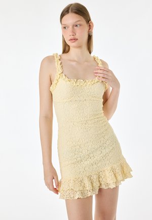 Jeune femme debout regardant sur le côté, vêtue d'une courte robe en dentelle jaune sans manches avec des bretelles et un ourlet à volants, sur fond uni.