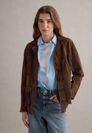 Vrouw draagt een bruine suède jas met franjes over een blauw gestreept overhemd en wit T-shirt, gecombineerd met hooggesneden blauwe spijkerbroek.