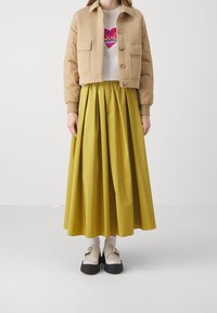 Khaki bomber jacket z dwoma przednimi kieszeniami, biała koszulka z grafiką oraz plisowana �żółta maks spódnica w połączeniu z masywnymi czarno-beżowymi butami.