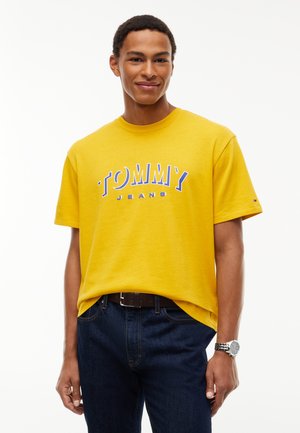 Νέος άνδρας με κίτρινο t-shirt Tommy Jeans και σκούρο μπλε τζιν, με μαύρη ζώνη και ασημένιο ρολόι χειρός, που στέκεται μπροστά σε λευκό φόντο.