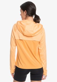 Huvtröja i atletisk stil i ljus orange tyg, med en ventilerande design, långa ärmar och en texturerad bakpanel.