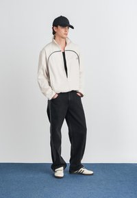 Mladý muž stojí s rukami v vreckách, oblečený v béžovej bundě na zips, čiernej čiapke, vo voľných čiernych džínsoch a bielych teniskách na modrom koberci.