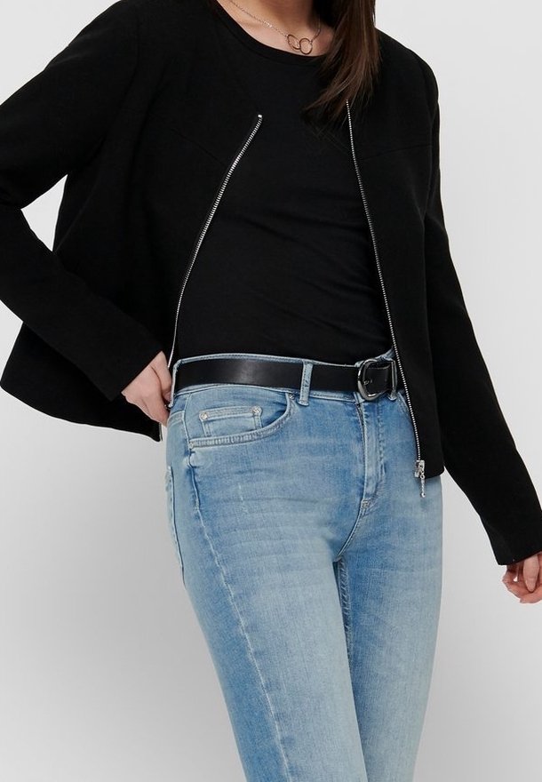 Veste noire courte avec fermeture éclair sur le devant et manches longues, portée sur une chemise noire. Associée à un jean bleu clair et une ceinture noire.