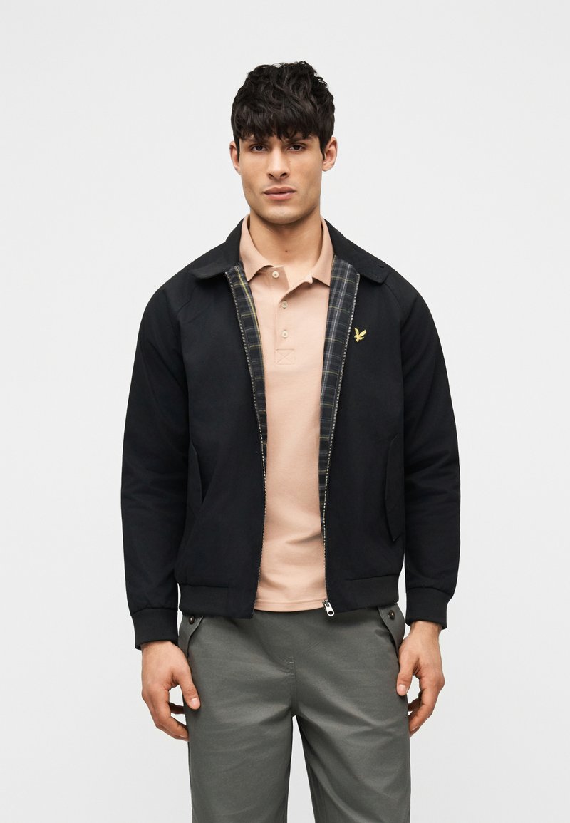 Άνδρας που φοράει μαύρο μπουφάν με καρό φόδρα πάνω από μπεζ πόλο πουκάμισο και γκρι παντελόνι, στέκεται μπροστά σε ένα απλό φωτεινό φόντο.