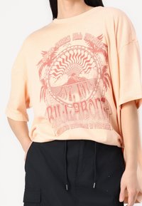 Camiseta de gran tamaño en color durazno, fabricada en algodón suave, que presenta un gran estampado gráfico de un sol y montañas en color coral, con mangas relajadas y cuello redondo.
