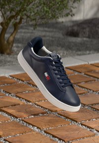 Baskets en cuir bleu foncé avec une semelle en caoutchouc blanc, présentant un petit patch logo rouge, blanc et bleu sur le côté. Lacets plats, design minimaliste.