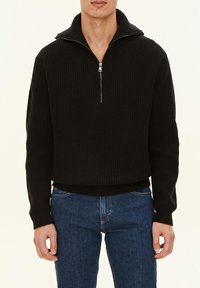 Svart stickad tröja med hög krage och halvdragkedja, med en texturerad ribbad design och en kort passform. Bärs tillsammans med blå jeans.