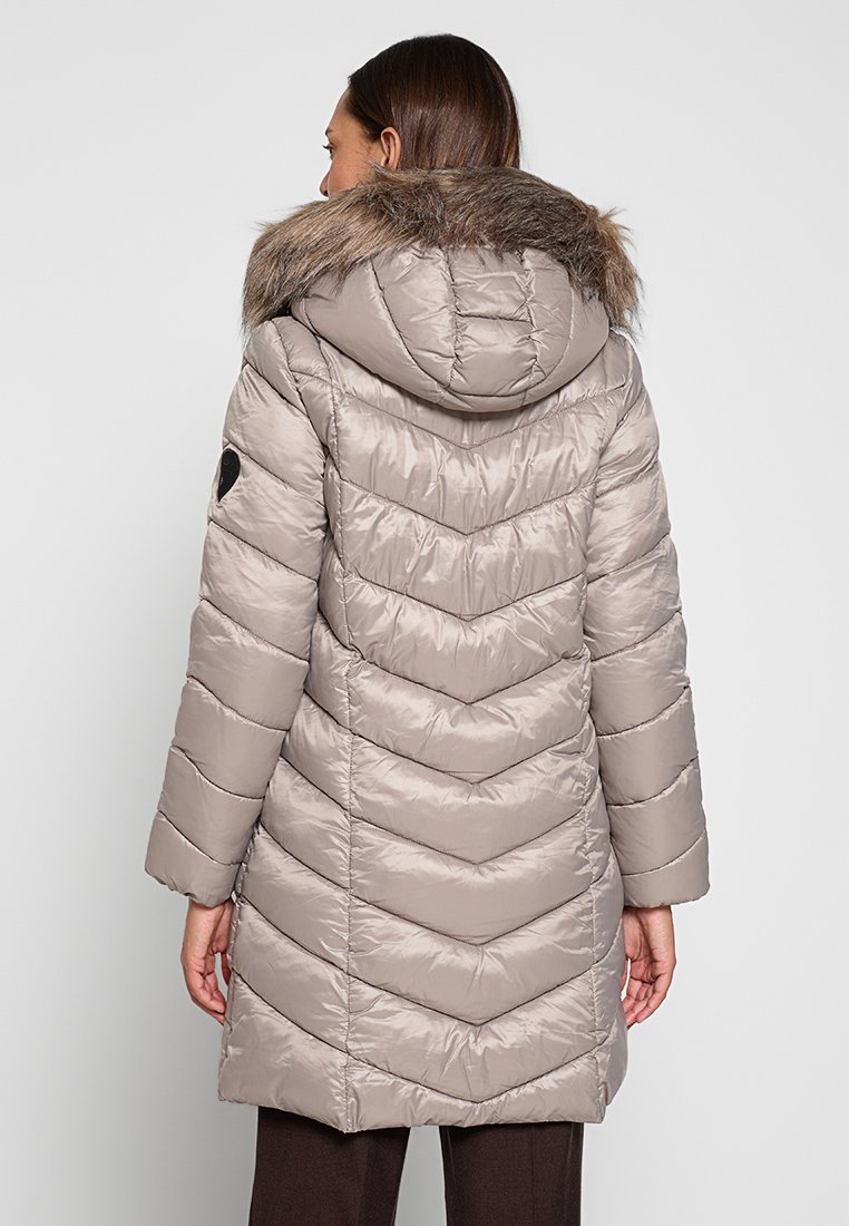 Donna che indossa un lungo cappotto invernale trapuntato di un beige chiaro con cappuccio rifinito in pelliccia, vista da dietro su uno sfondo semplice.
