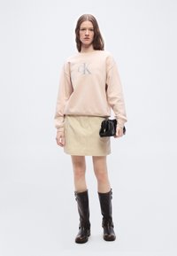 Γυναίκα που φορεί μπεζ φούτερ Calvin Klein, φούστα καφέ ανοιχτού χρώματος, σκούρα καφέ μπότες μέχρι το γόνατο, κρατώντας μαύρο τσαντάκι ruched σε λευκό φόντο.