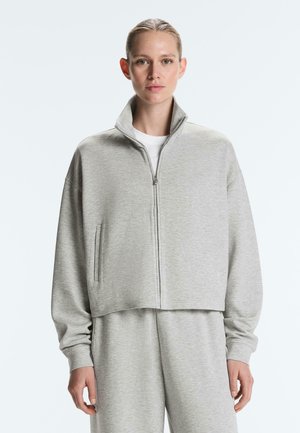 Vrouw draagt een lichtgrijze zip-up sporttrui en bijpassende joggingbroek, staand tegen een effen lichte achtergrond met een neutrale uitdrukking.