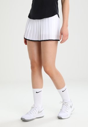 Osoba nosící bílou plisovanou tenisovou sukni, černý top, bílé ponožky Nike a bílé tenisové boty Nike stojí na jednobarevném pozadí.