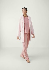 Naine kõnnib paljajalu pehmes roosas vabalajuriietuses, mis sisaldab hommikumantlit, pika varrukaga tuba ja pükse, tavalise heleda taustaga.