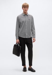 Melnbalts svītrains garām piedurknēm krekls ar pogām pie apkakles, kombinēts ar melnām biksēm un loafers; tur rokā melnu ādas somu.