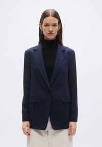 Granatowa marynarka z wyraźnie zarysowanymi ramionami, zapinana na jeden guzik, z dwoma przednimi kieszeniami i gładką fakturą, noszona na czarnej golfie.