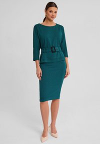 Turkusowa sukienka w wzór houndstooth, z peplum w talii i rękawami o długości trzy czwarte, z szerokim czarnym paskiem i sylwetką ołówkową.