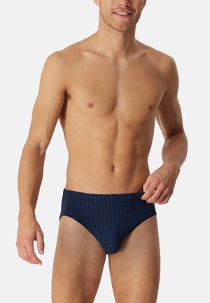 Ein oberkörperfreier Mann lächelt, trägt marineblaue Unterhosen mit einem dezenten Karomuster und steht vor einem schlichten weißen Hintergrund.