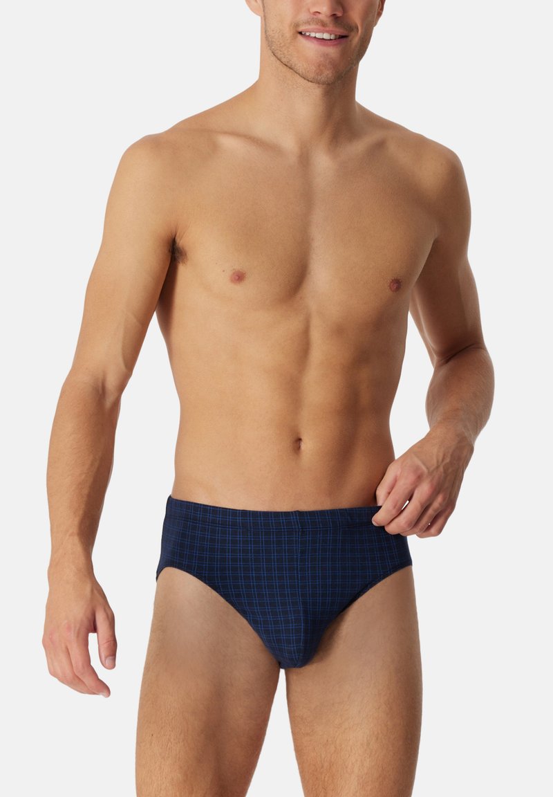 Ein oberkörperfreier Mann lächelt, trägt marineblaue Unterhosen mit einem dezenten Karomuster und steht vor einem schlichten weißen Hintergrund.