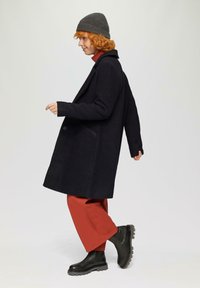 Lange donkere visgraatjas met een kraag, zijzakken en knopen sluiting, gecombineerd met oranje wijde broekspijpen en zwarte enkellaarsjes.